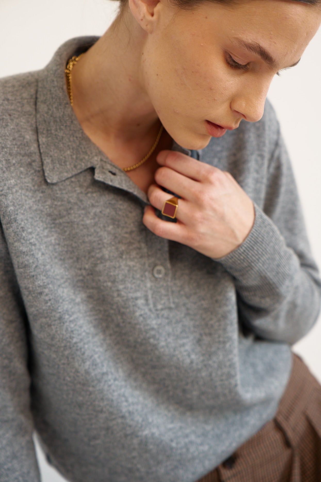A close-up lifestyle shot of a model wearing a gray knit polo sweater, a gold chain necklace, and a gold ring with a red stone.
