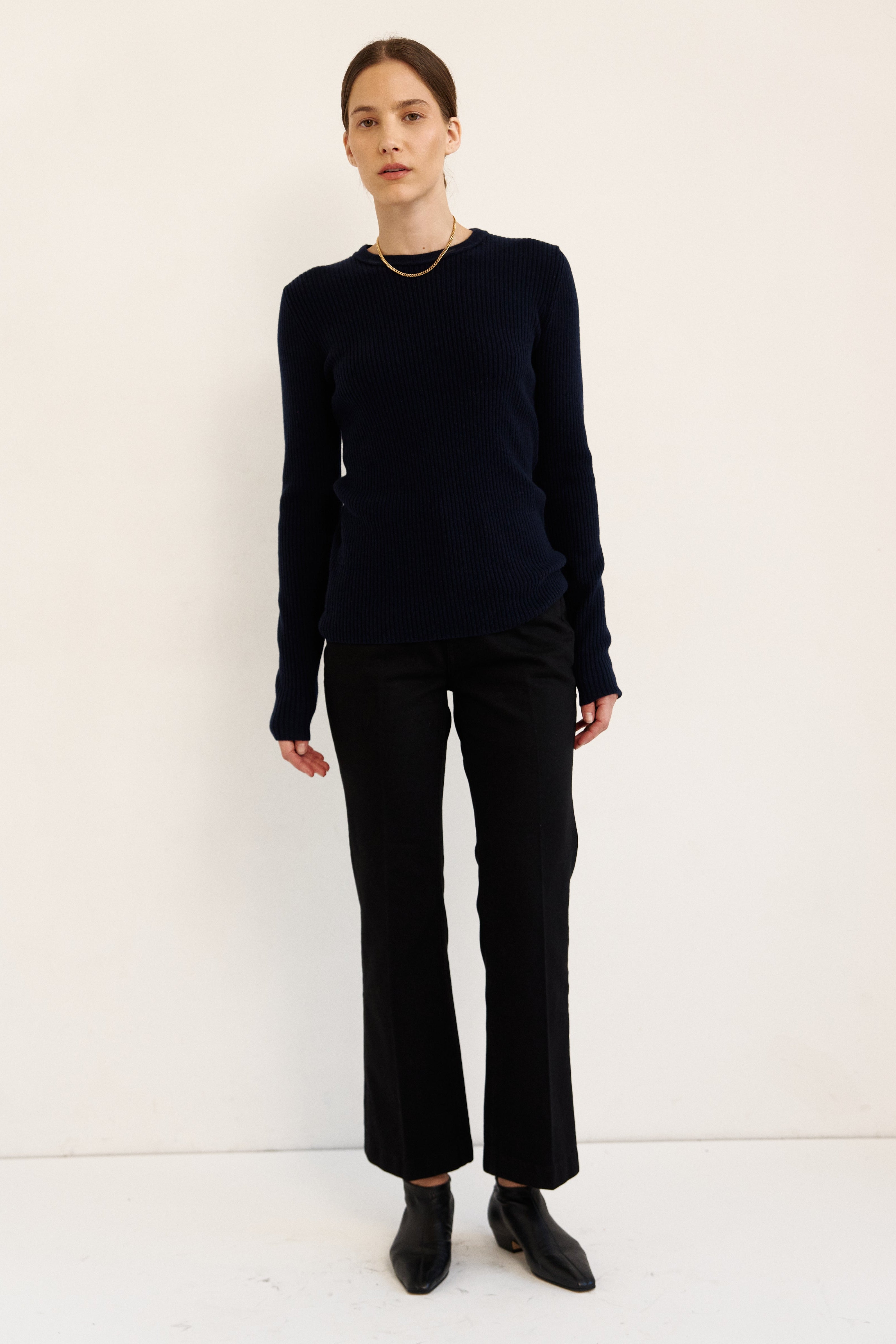 A woman modeling a dark navy long-sleeve knit top and black wide-leg trousers with black ankle boots, standing against a plain off-white studio background.