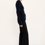 A woman stands in profile wearing a dark navy long-sleeve knit top, black wide-leg cropped trousers, and black shoes against a clean white studio background.