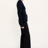 A woman stands in profile wearing a dark navy long-sleeve knit top, black wide-leg cropped trousers, and black shoes against a clean white studio background.