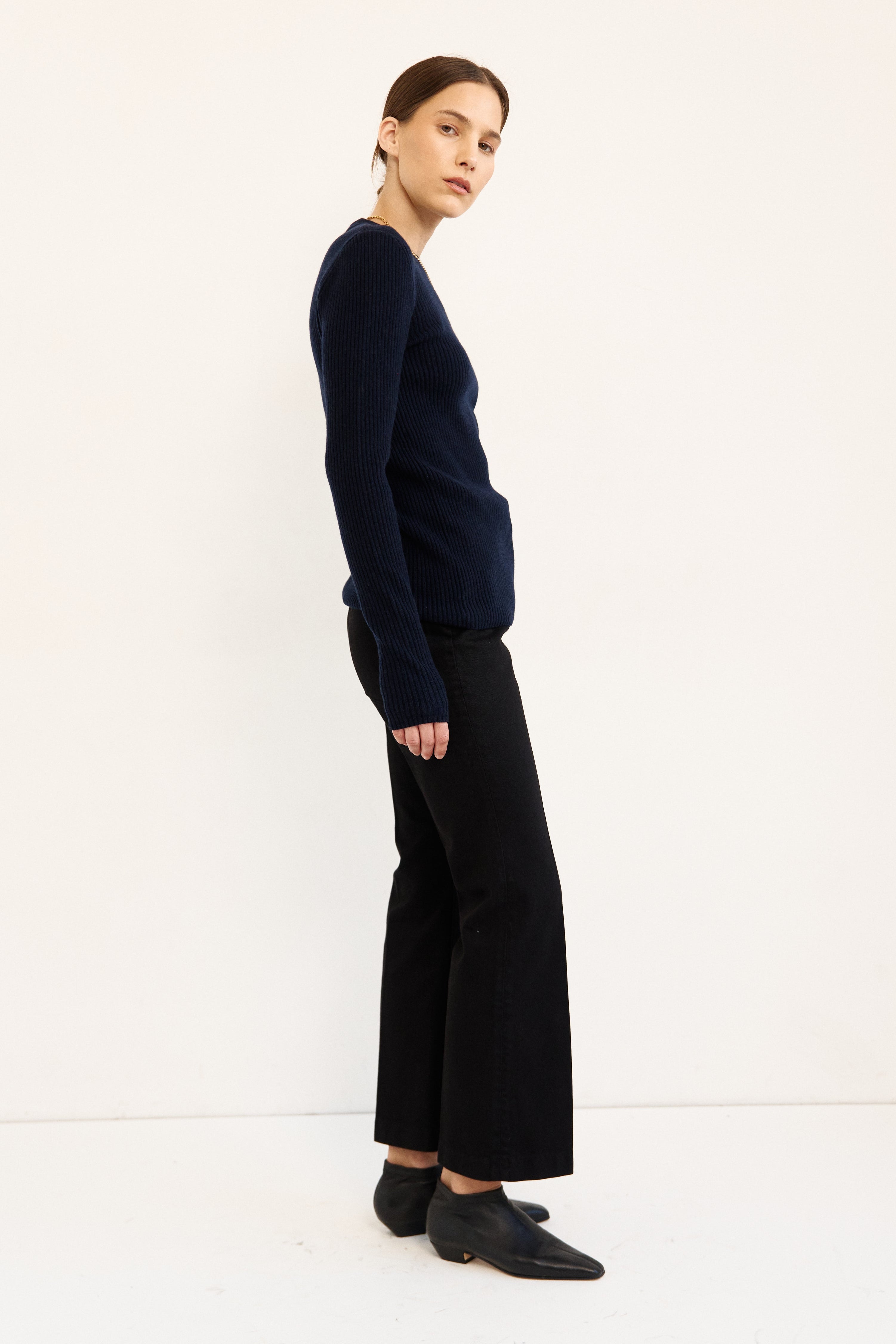 A woman stands in profile wearing a dark navy long-sleeve knit top, black wide-leg cropped trousers, and black shoes against a clean white studio background.