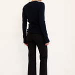 A woman stands in profile against a plain white studio background, wearing a dark navy long-sleeve knit top, black cropped pants, and black pointed shoes.