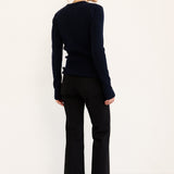 A woman stands in profile against a plain white studio background, wearing a dark navy long-sleeve knit top, black cropped pants, and black pointed shoes.