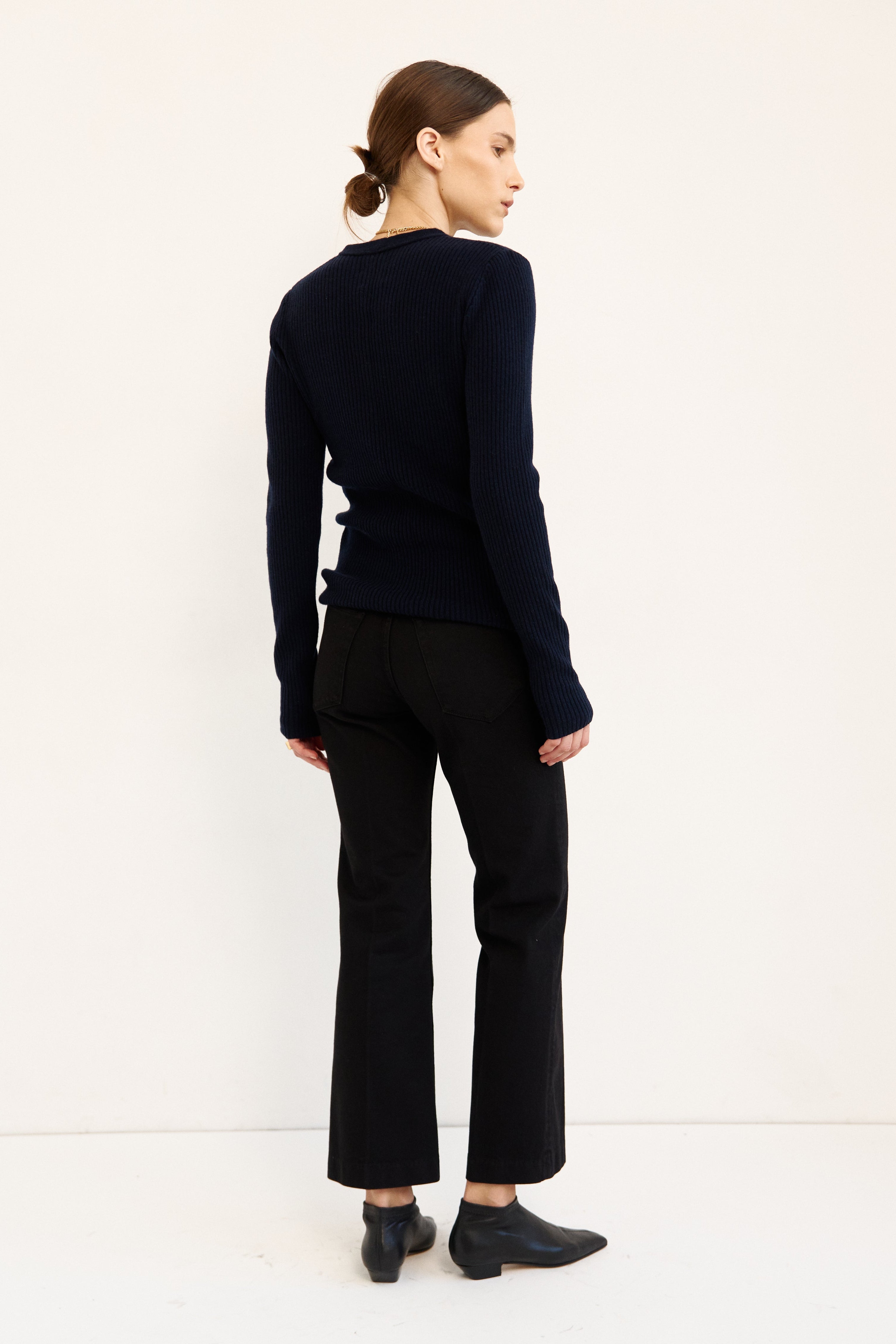 A woman stands in profile against a plain white studio background, wearing a dark navy long-sleeve knit top, black cropped pants, and black pointed shoes.