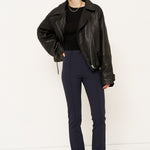 A model stands against a plain white studio background wearing a black leather moto jacket, a black top, navy blue trousers, and black pointed-toe boots.
