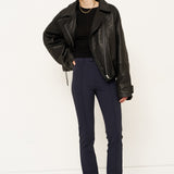 A model stands against a plain white studio background wearing a black leather moto jacket, a black top, navy blue trousers, and black pointed-toe boots.