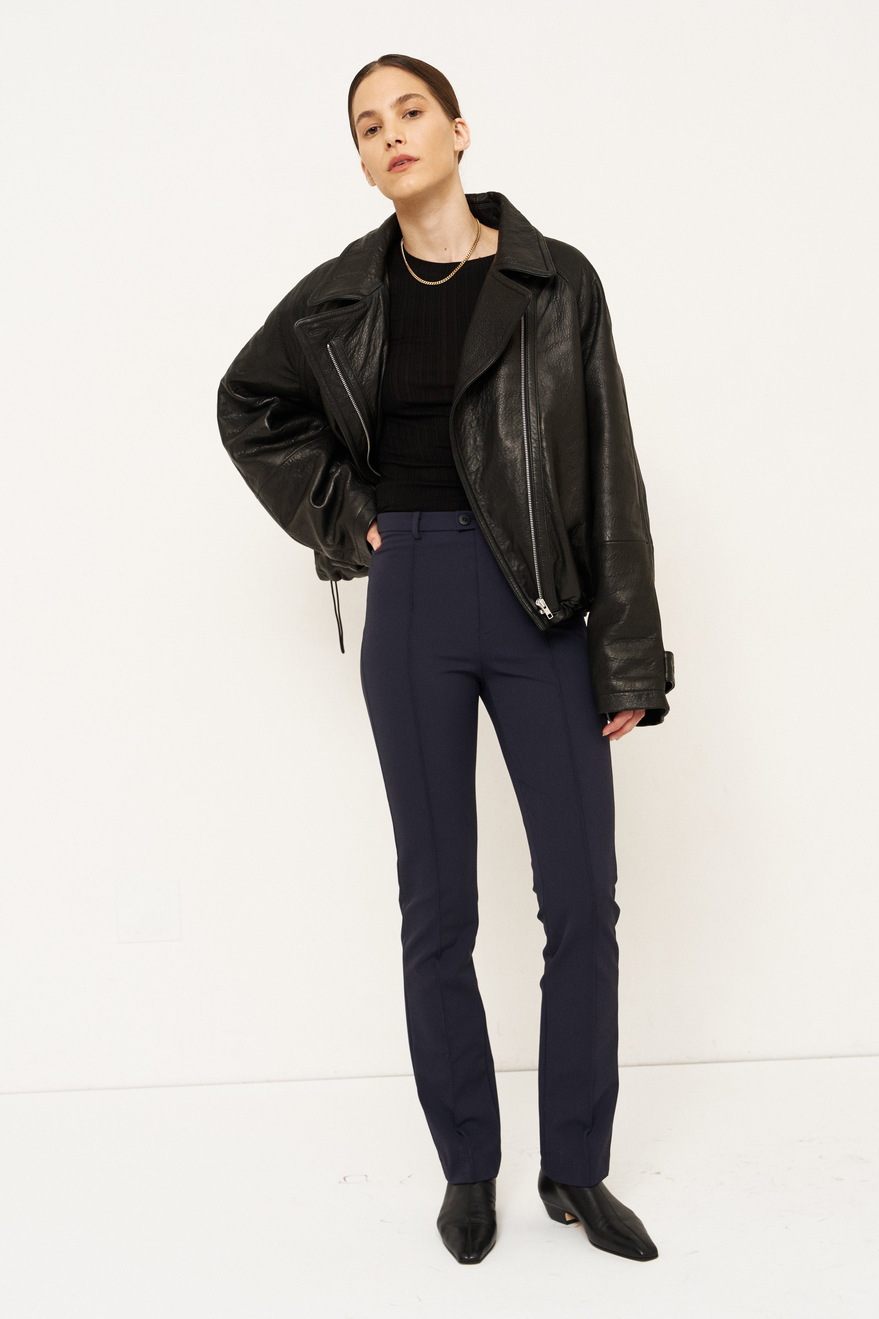 A model stands against a plain white studio background wearing a black leather moto jacket, a black top, navy blue trousers, and black pointed-toe boots.