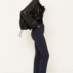A woman stands in profile wearing a black leather jacket, black top, dark navy trousers, and black pointed shoes against a plain white studio background.