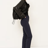 A woman stands in profile wearing a black leather jacket, black top, dark navy trousers, and black pointed shoes against a plain white studio background.
