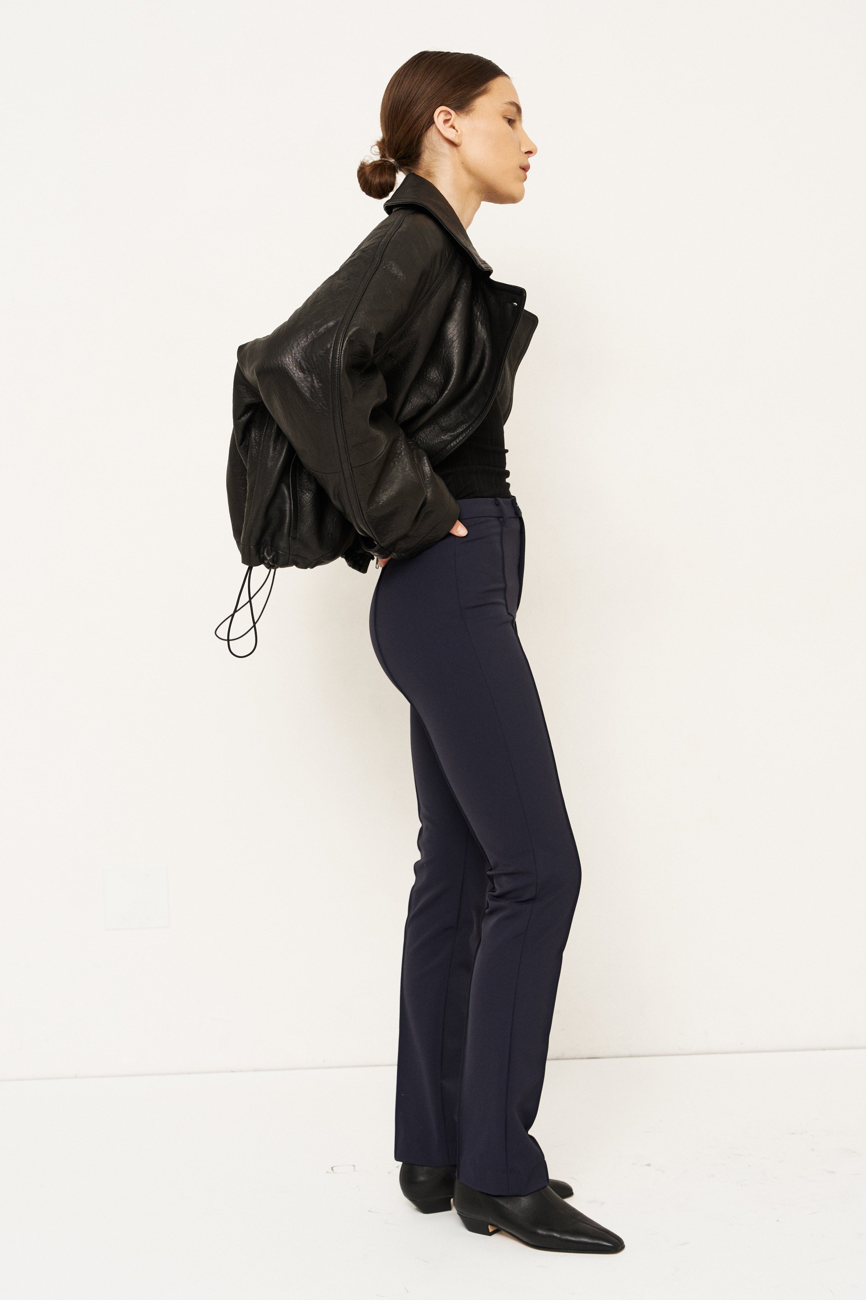 A woman stands in profile wearing a black leather jacket, black top, dark navy trousers, and black pointed shoes against a plain white studio background.