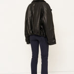 A model viewed from the back wearing a black leather jacket, navy blue trousers, and black shoes, standing against a plain white studio background.