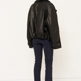 A model viewed from the back wearing a black leather jacket, navy blue trousers, and black shoes, standing against a plain white studio background.