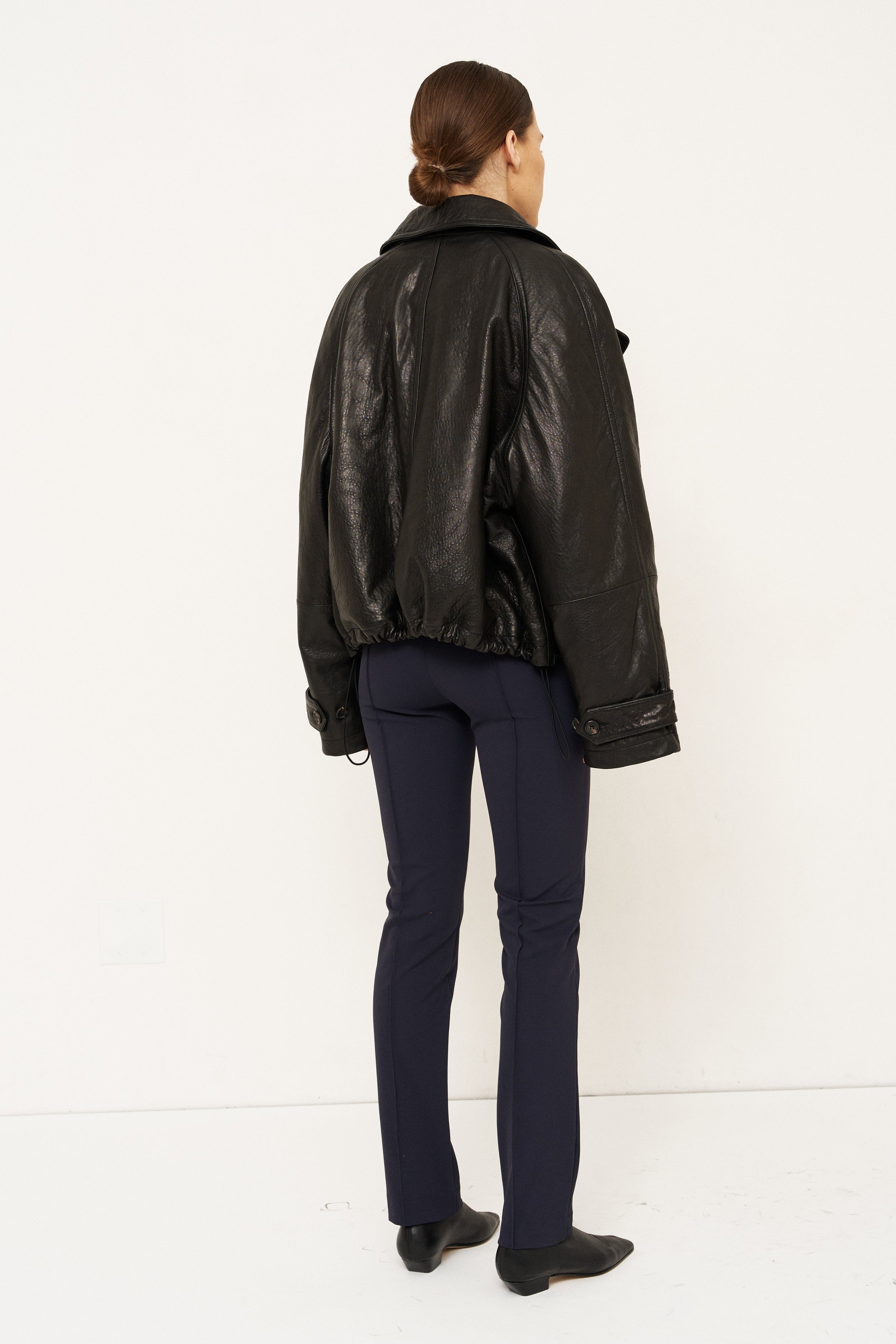 A model viewed from the back wearing a black leather jacket, navy blue trousers, and black shoes, standing against a plain white studio background.