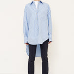 A model stands in profile wearing a light blue and white vertically striped button-up shirt, dark navy trousers, and black boots against a plain white studio background.