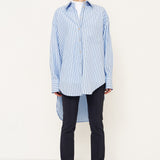 A model stands in profile wearing a light blue and white vertically striped button-up shirt, dark navy trousers, and black boots against a plain white studio background.