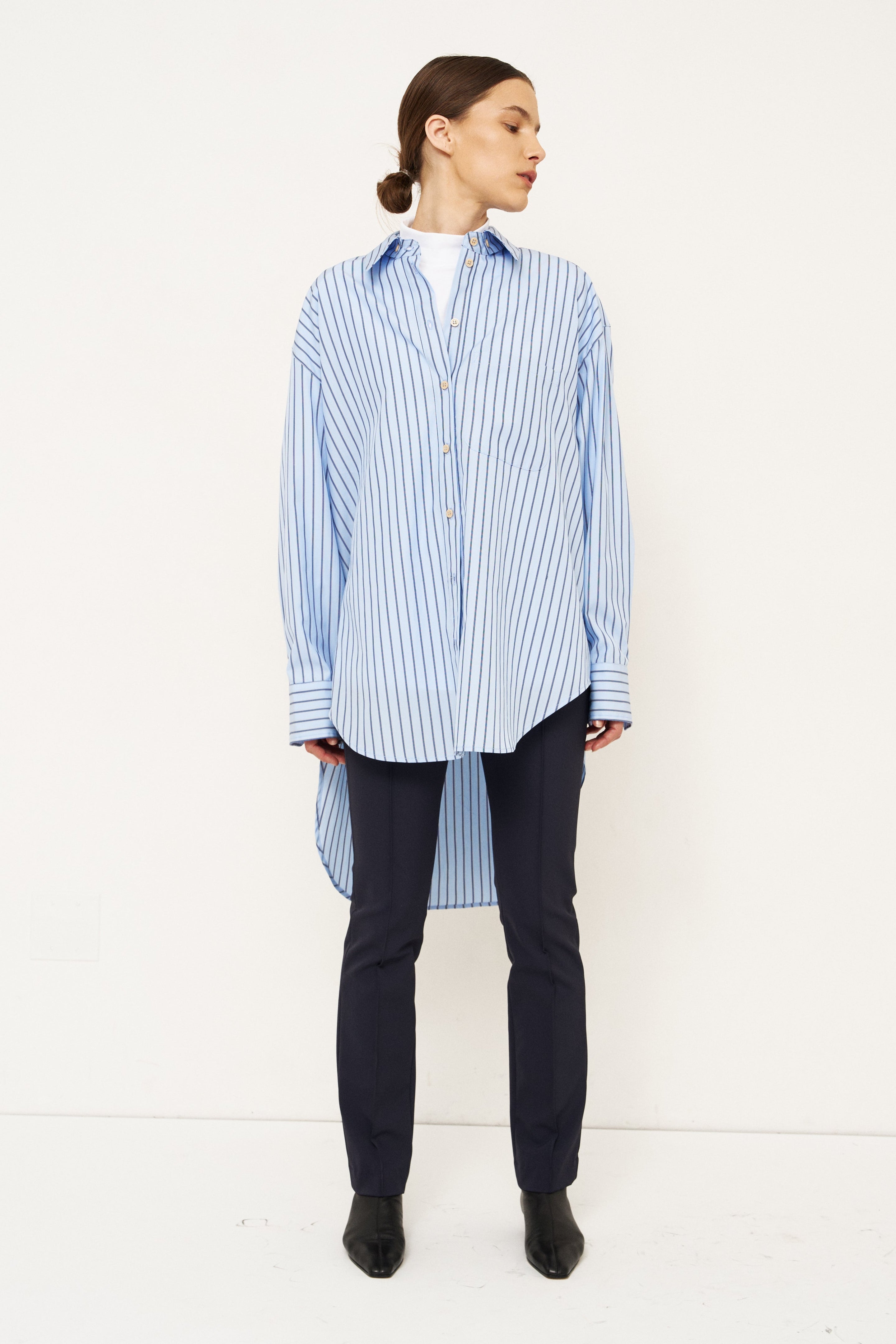 A model stands in profile wearing a light blue and white vertically striped button-up shirt, dark navy trousers, and black boots against a plain white studio background.
