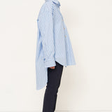 A model shown in profile wearing a light blue and white vertically striped button-down shirt, dark navy trousers, and black shoes, standing against a clean white studio background.