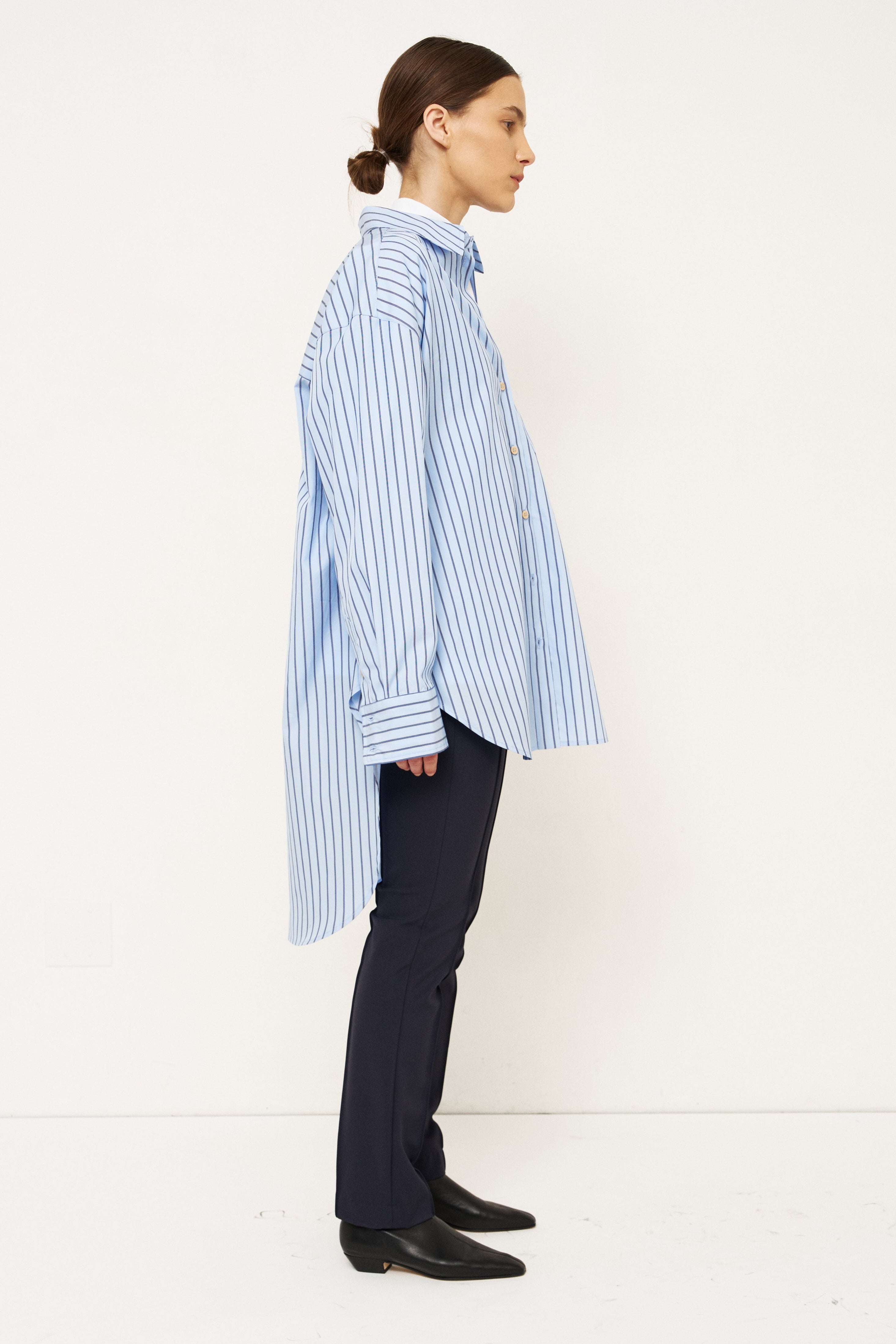 A model shown in profile wearing a light blue and white vertically striped button-down shirt, dark navy trousers, and black shoes, standing against a clean white studio background.