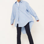 A woman modeling an oversized light blue and white vertically striped button-down shirt with a high-low hem, paired with dark trousers and black shoes, against a plain white studio background.