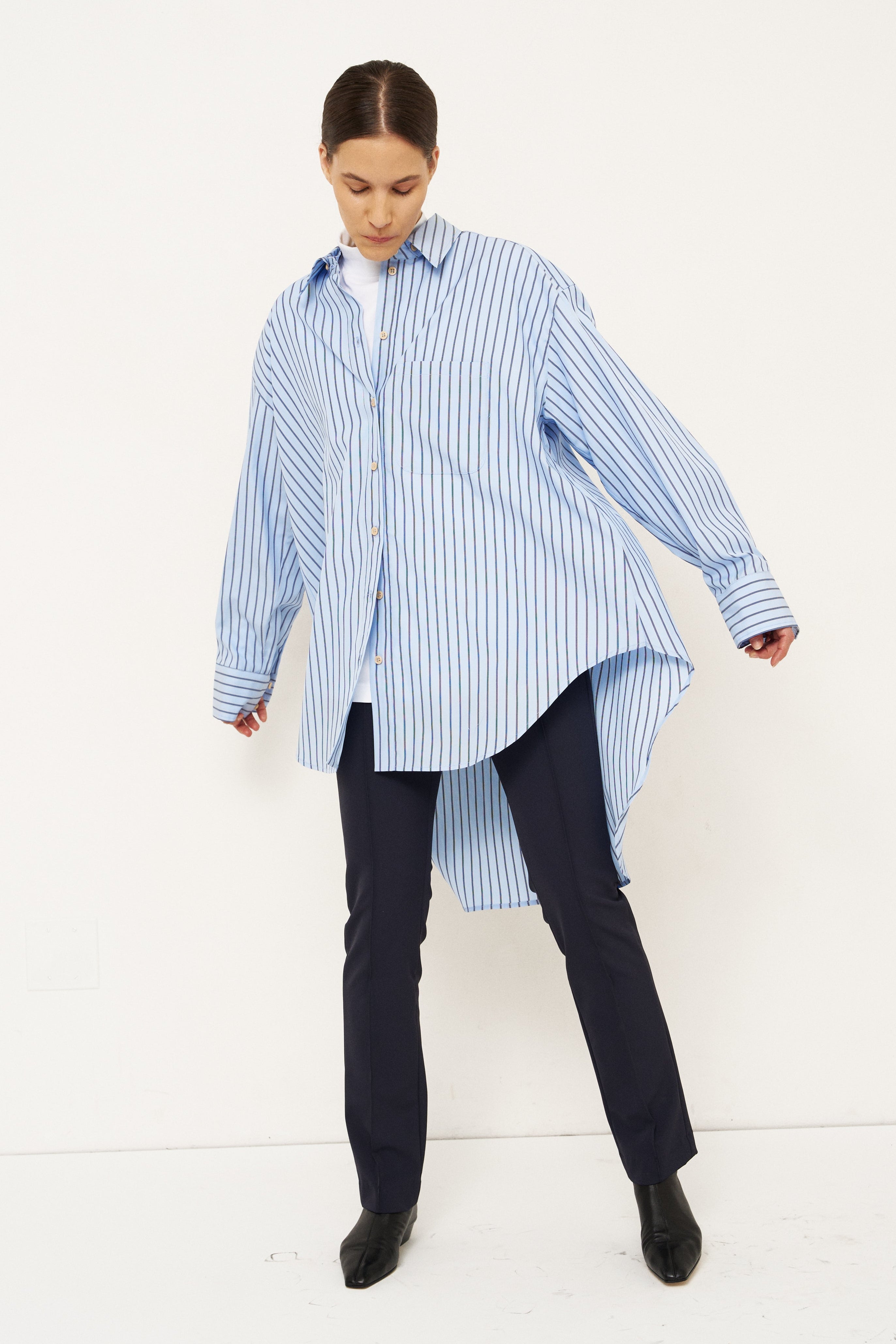 A woman modeling an oversized light blue and white vertically striped button-down shirt with a high-low hem, paired with dark trousers and black shoes, against a plain white studio background.