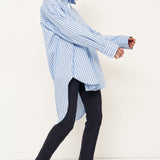 A woman modeling an oversized light blue and white vertically striped button-down shirt, paired with dark trousers and black shoes, captured in a side-profile pose against a clean white studio background.