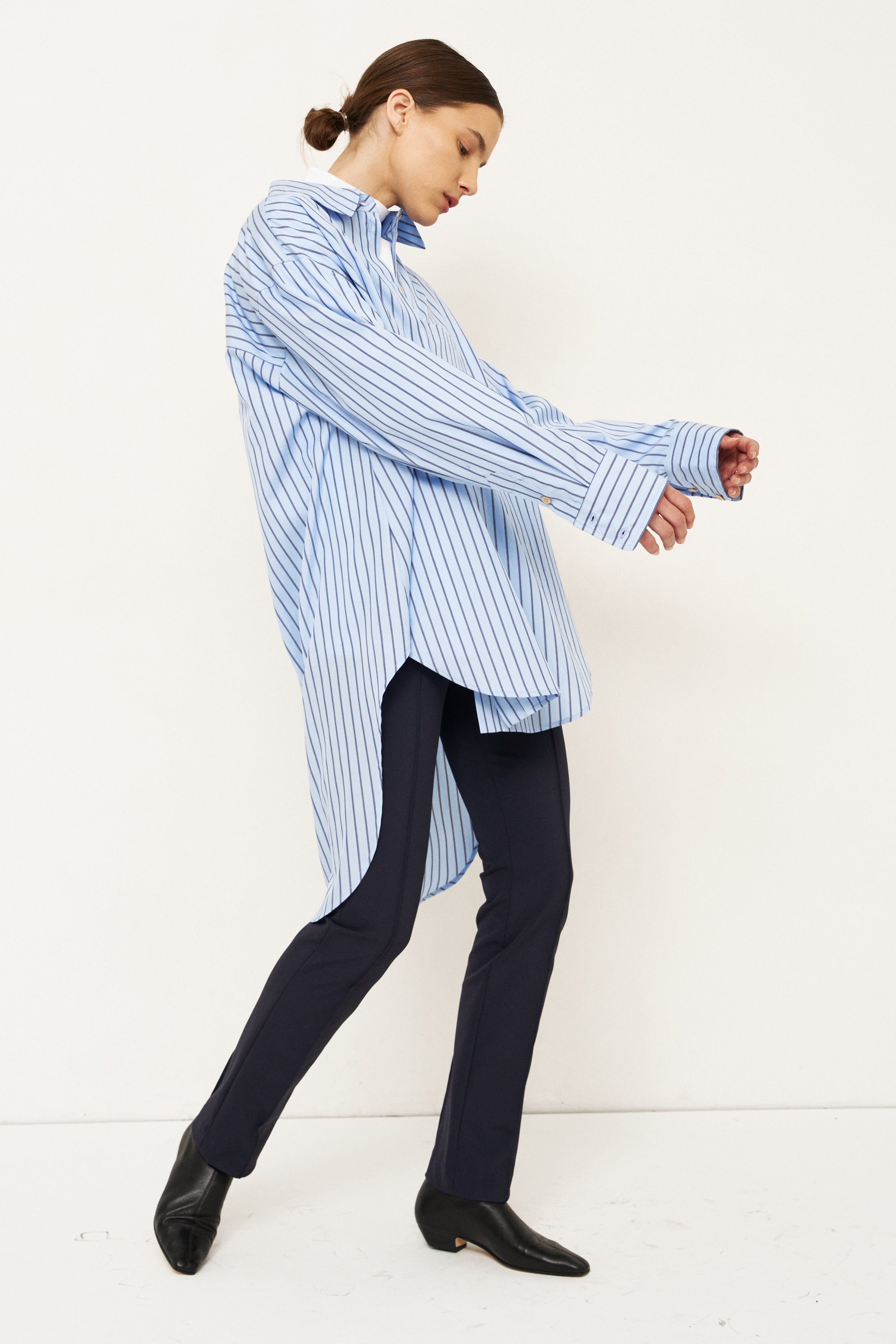 A woman modeling an oversized light blue and white vertically striped button-down shirt, paired with dark trousers and black shoes, captured in a side-profile pose against a clean white studio background.