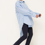 A model wearing a light blue and white vertically striped oversized button-down shirt, dark navy trousers, and black pointed-toe shoes, captured in a dynamic pose against a clean white studio background.