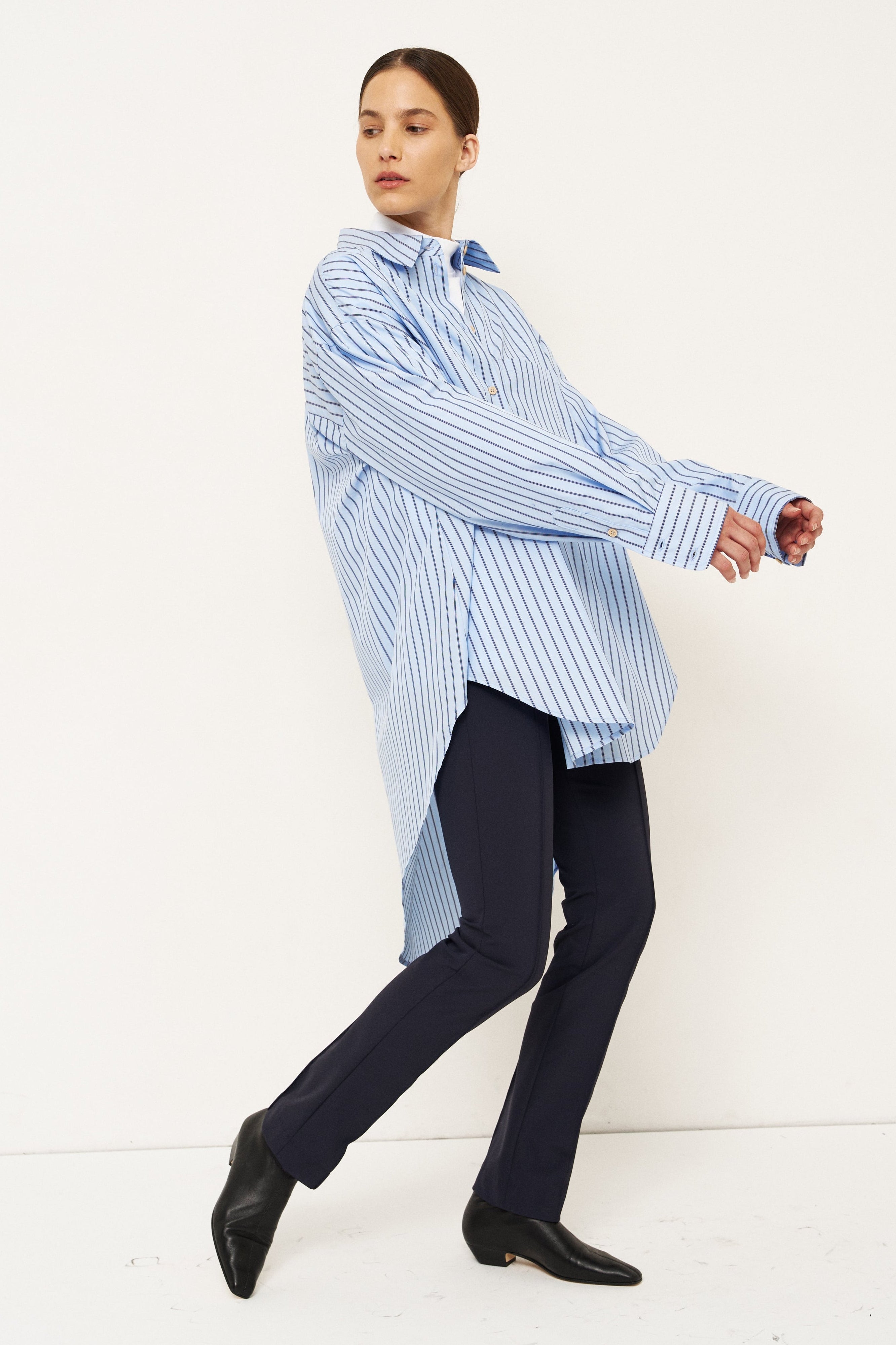 A model wearing a light blue and white vertically striped oversized button-down shirt, dark navy trousers, and black pointed-toe shoes, captured in a dynamic pose against a clean white studio background.