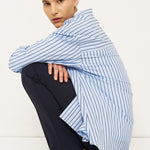 A model crouches in a studio setting wearing a light blue and white vertically striped shirt draped over dark navy trousers and black leather shoes.