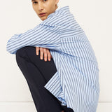 A model crouches in a studio setting wearing a light blue and white vertically striped shirt draped over dark navy trousers and black leather shoes.
