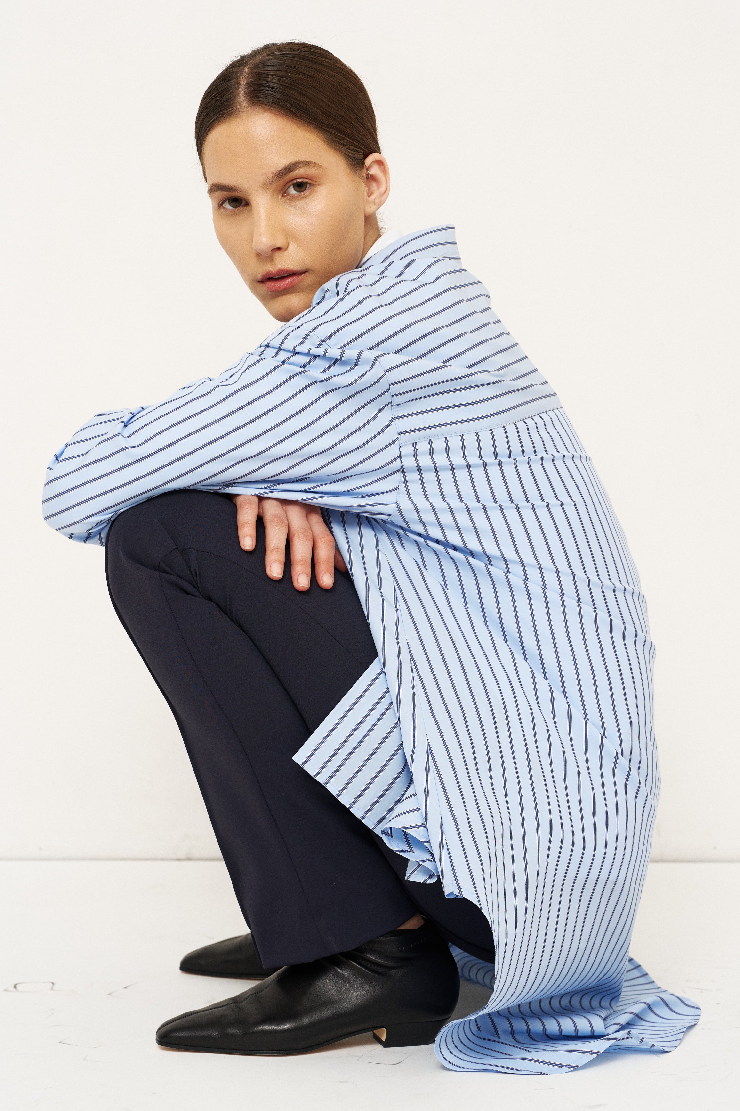 A model crouches in a studio setting wearing a light blue and white vertically striped shirt draped over dark navy trousers and black leather shoes.