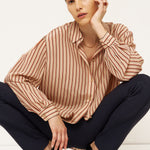 A model crouching in a studio setting, wearing a beige and brown vertically striped long-sleeve blouse, dark navy trousers, and black pointed-toe heels with gold heels.