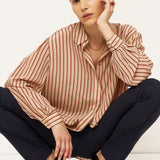 A model crouching in a studio setting, wearing a beige and brown vertically striped long-sleeve blouse, dark navy trousers, and black pointed-toe heels with gold heels.