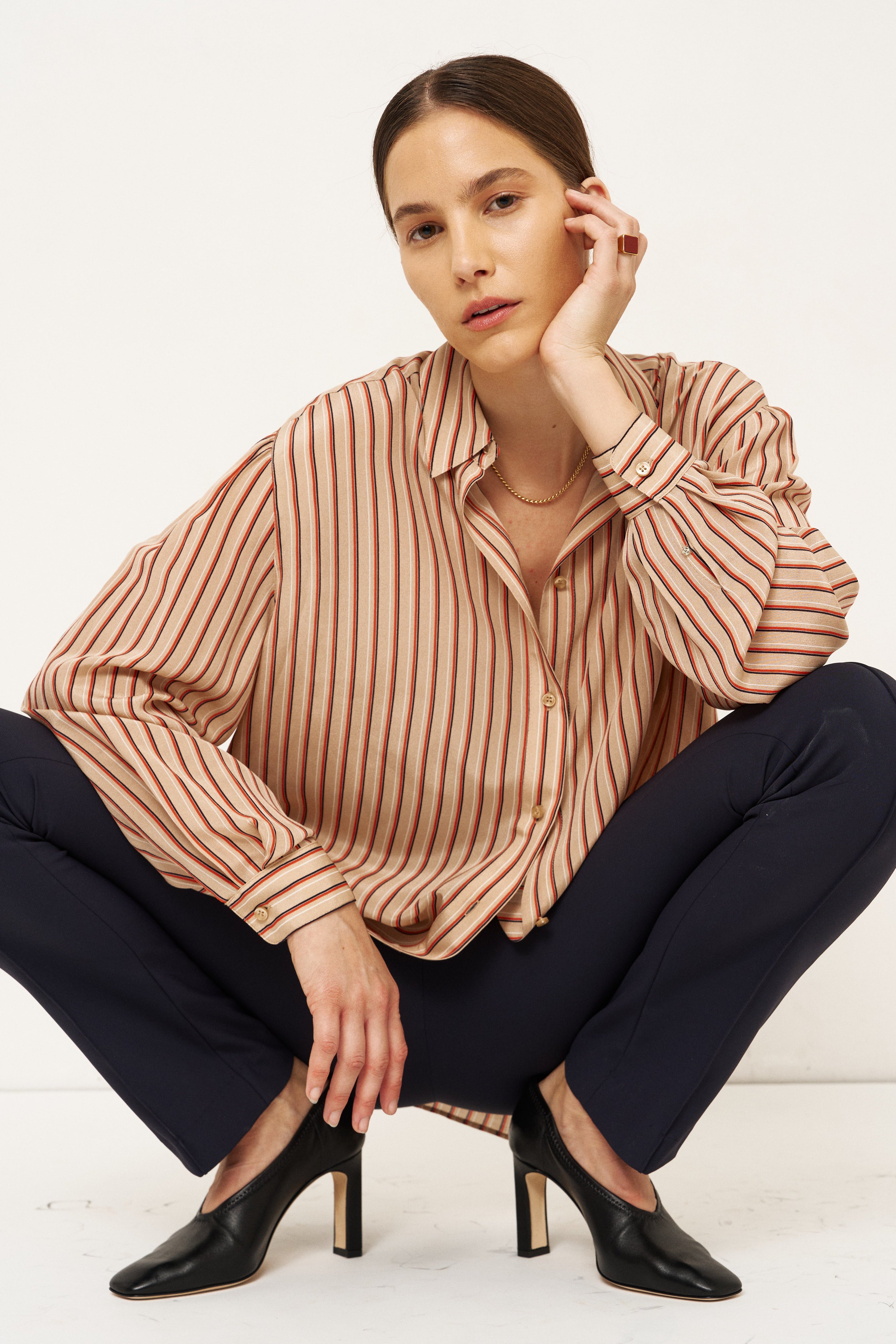 A model crouching in a studio setting, wearing a beige and brown vertically striped long-sleeve blouse, dark navy trousers, and black pointed-toe heels with gold heels.