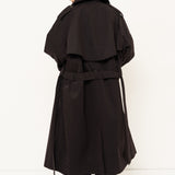 A model viewed from behind wearing a long black trench coat with a belted waist, dark trousers, and red shoes, standing against a plain white studio background.