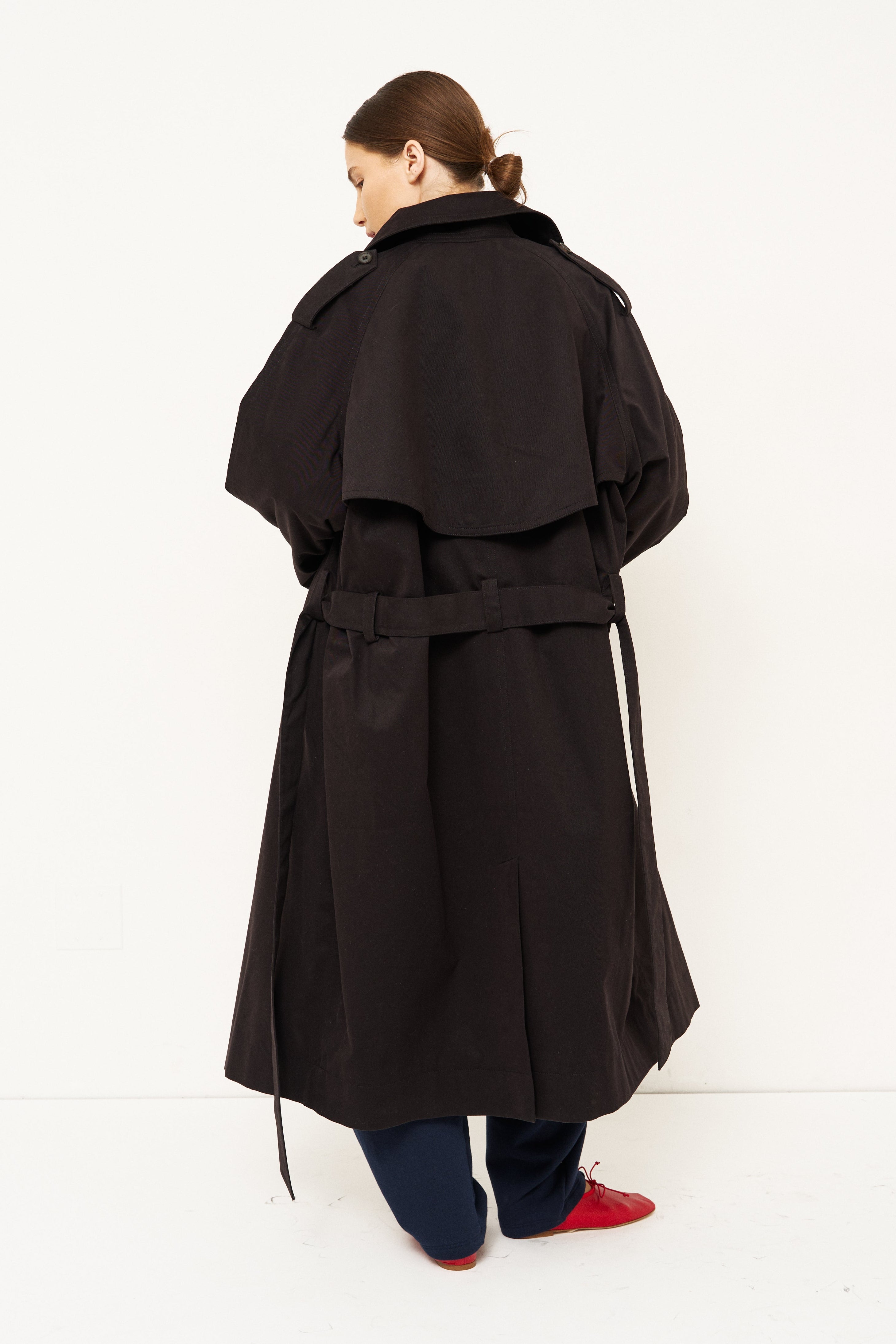 A model viewed from behind wearing a long black trench coat with a belted waist, dark trousers, and red shoes, standing against a plain white studio background.