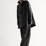 A model wearing a black oversized long-sleeve shirt and matching wide-leg trousers, styled with black pointed-toe heels, standing against a plain white studio background.