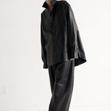 A model wearing a black oversized long-sleeve shirt and matching wide-leg trousers, styled with black pointed-toe heels, standing against a plain white studio background.