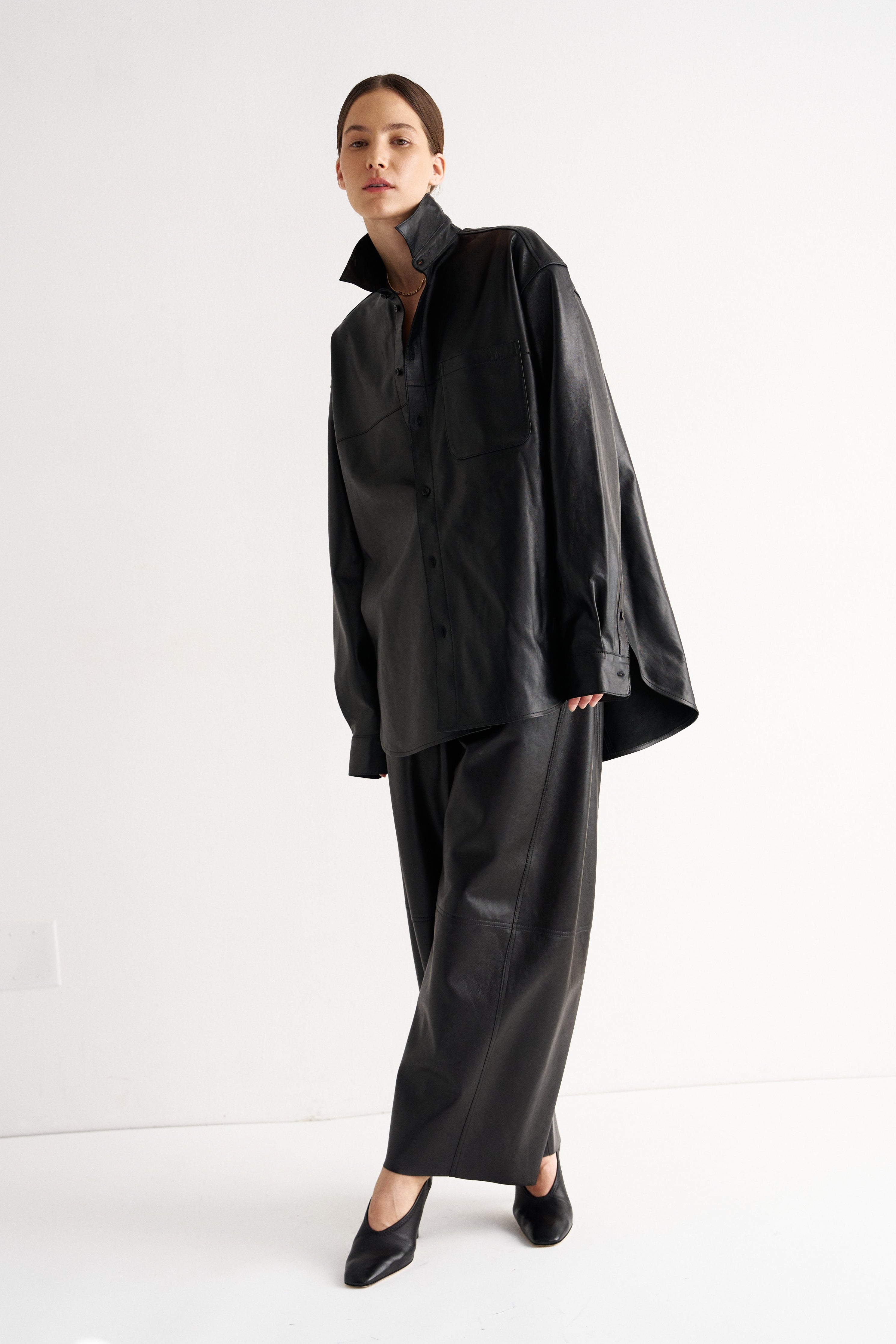 A model wearing a black oversized long-sleeve shirt and matching wide-leg trousers, styled with black pointed-toe heels, standing against a plain white studio background.
