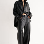 A model stands in a studio wearing a black oversized collared shirt jacket, matching wide-leg leather-look pants, and black pointed heels against a clean white background.