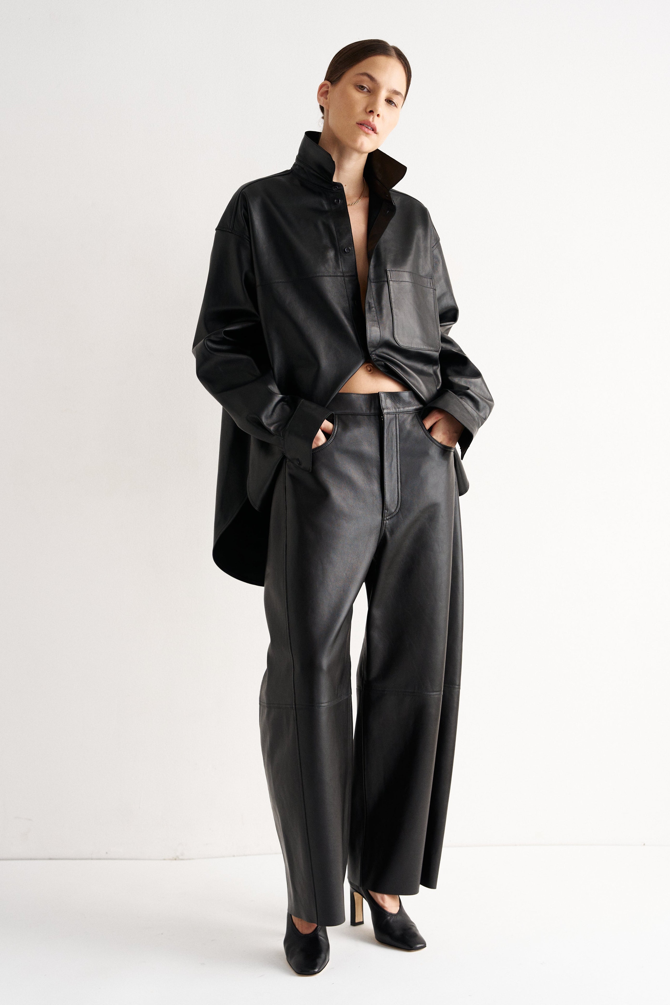 A model stands in a studio wearing a black oversized collared shirt jacket, matching wide-leg leather-look pants, and black pointed heels against a clean white background.