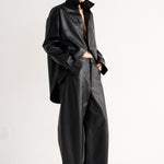 A model stands in a minimalist studio setting wearing a black leather-like cropped jacket with a high collar, matching wide-leg pants, and black pointed shoes.