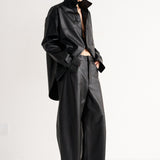 A model stands in a minimalist studio setting wearing a black leather-like cropped jacket with a high collar, matching wide-leg pants, and black pointed shoes.