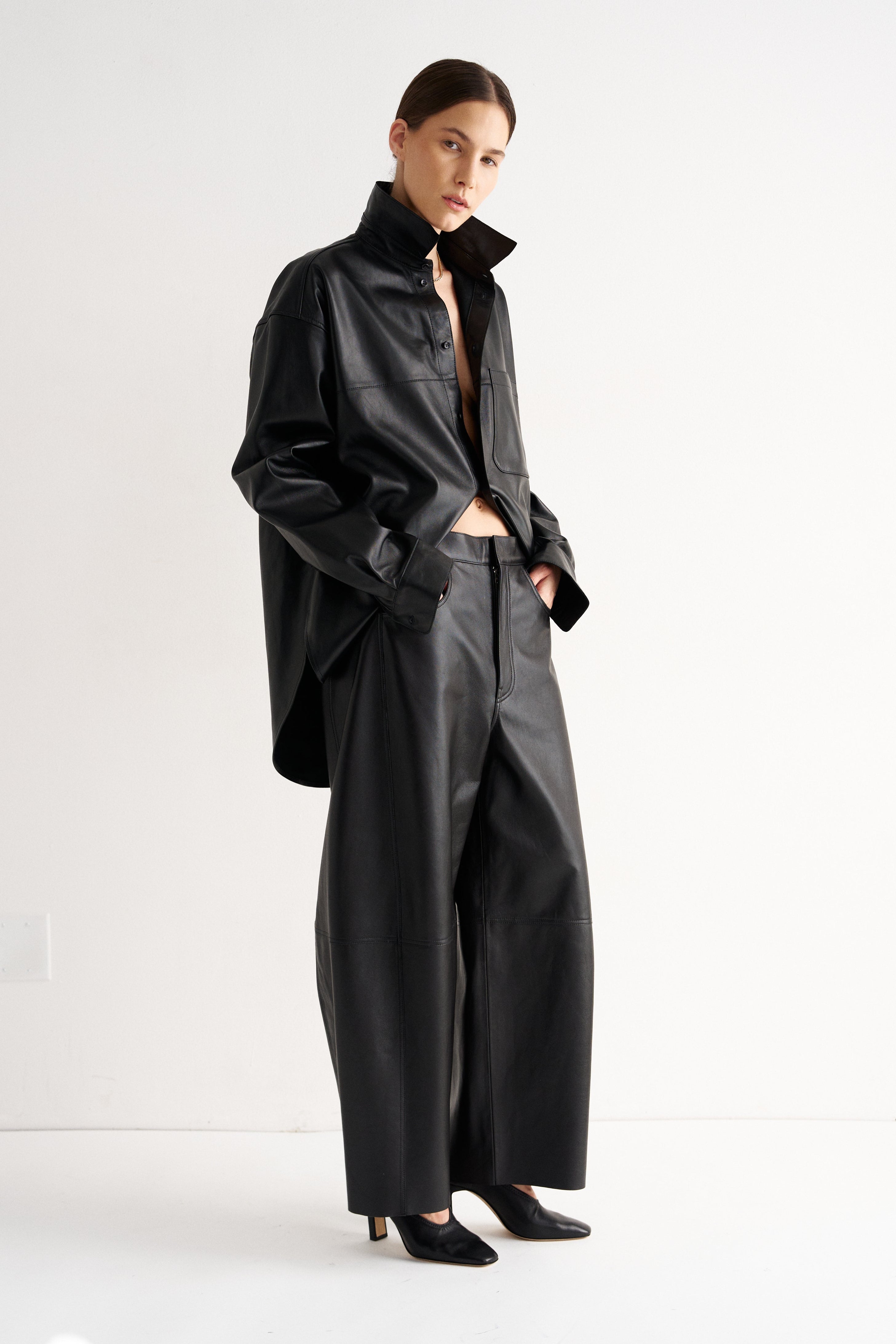A model stands in a minimalist studio setting wearing a black leather-like cropped jacket with a high collar, matching wide-leg pants, and black pointed shoes.