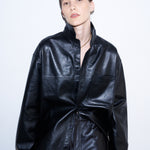 A model wearing a black faux-leather jumpsuit with a high collar and front button placket, styled with matching black trousers against a plain white studio background.