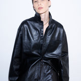 A model wearing a black faux-leather jumpsuit with a high collar and front button placket, styled with matching black trousers against a plain white studio background.
