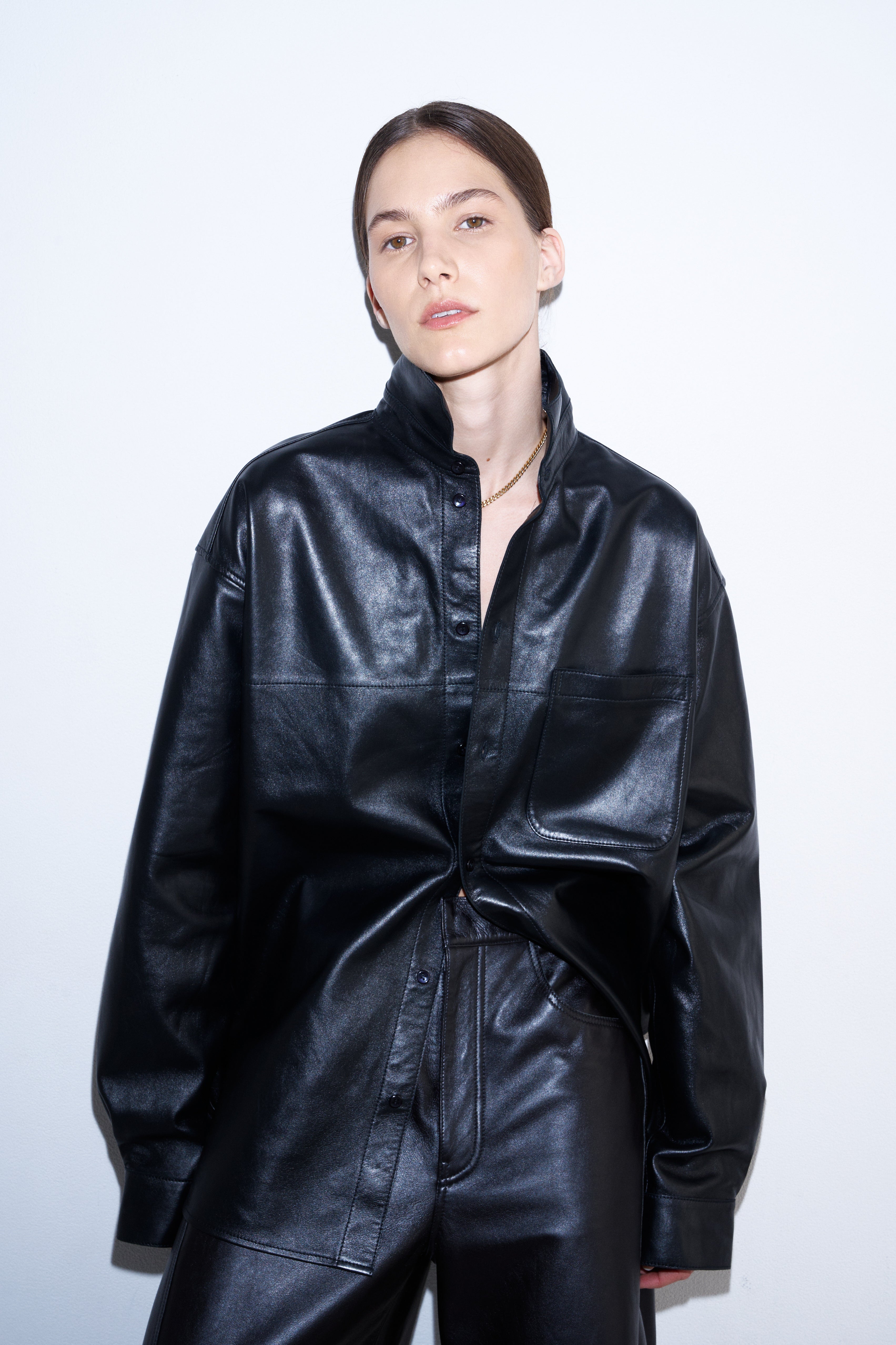 A model wearing a black faux-leather jumpsuit with a high collar and front button placket, styled with matching black trousers against a plain white studio background.