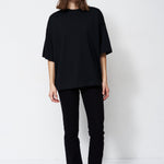 A model stands against a plain white studio background wearing a loose black short-sleeve top, black pants, and black pointed shoes.