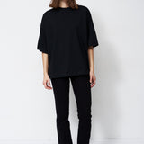 A model stands against a plain white studio background wearing a loose black short-sleeve top, black pants, and black pointed shoes.