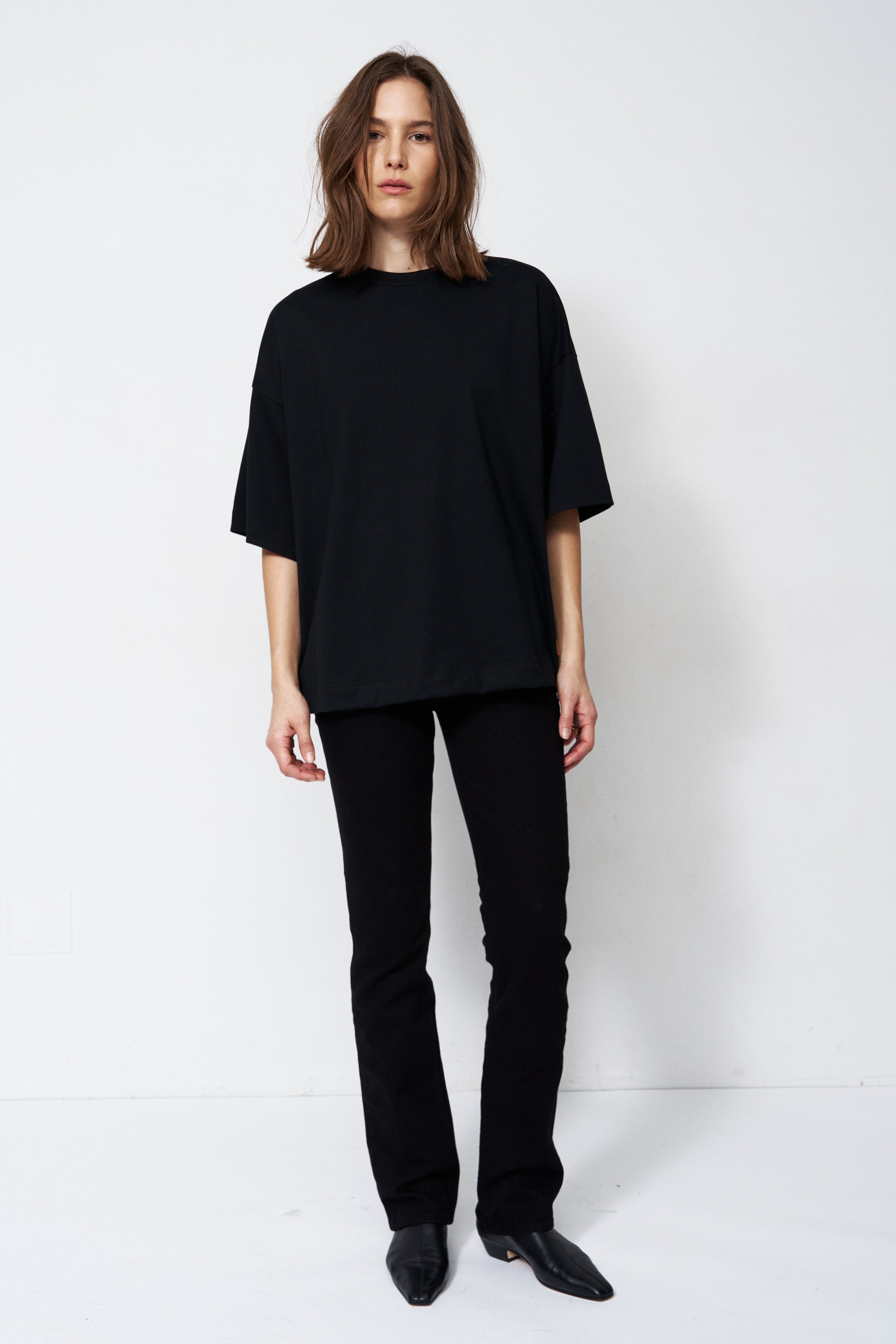 A model stands against a plain white studio background wearing a loose black short-sleeve top, black pants, and black pointed shoes.